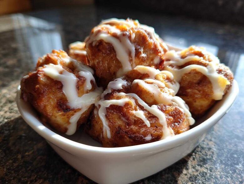 A white bowl filled with freshly baked, golden brown cinnamon roll bites drizzled generously with white icing.