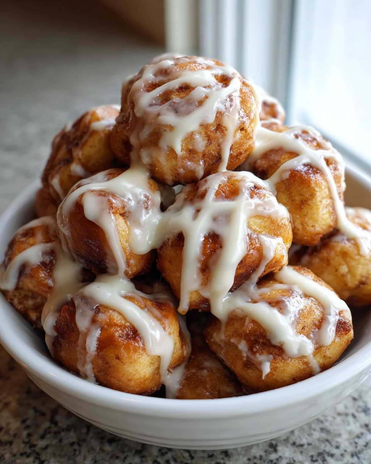 A white bowl overflowing with golden brown cinnamon roll bites drizzled generously with white cream cheese icing.