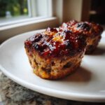 Close-up of a perfectly glazed mini meatloaf sitting on a white plate near a window.