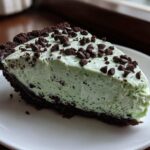 A close-up of a creamy, light green slice of grasshopper pie with a dark chocolate crust, topped with chocolate chips.