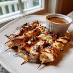 Four skewers of perfectly grilled chicken satay served on a plate next to a small bowl of peanut dipping sauce.