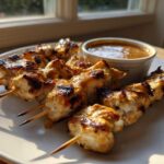 Close-up of perfectly grilled chicken satay skewers served on a white plate next to a small bowl of peanut dipping sauce.