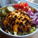 Close-up of a delicious cheeseburger bowl featuring seasoned ground beef, melted cheese sauce, lettuce, pickles, tomatoes, and red onion.