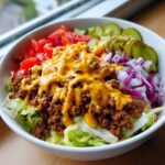 Close-up of a vibrant cheeseburger bowl featuring seasoned ground beef, melted cheese sauce, lettuce, tomatoes, red onion, and pickles.