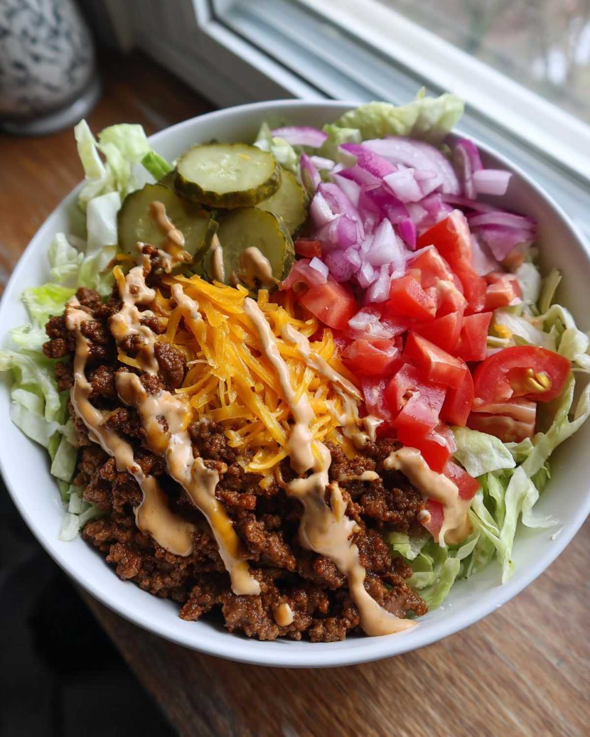 Close-up of a high-protein cheeseburger bowl featuring seasoned ground beef, shredded cheese, pickles, tomatoes, onions, and sauce over lettuce.
