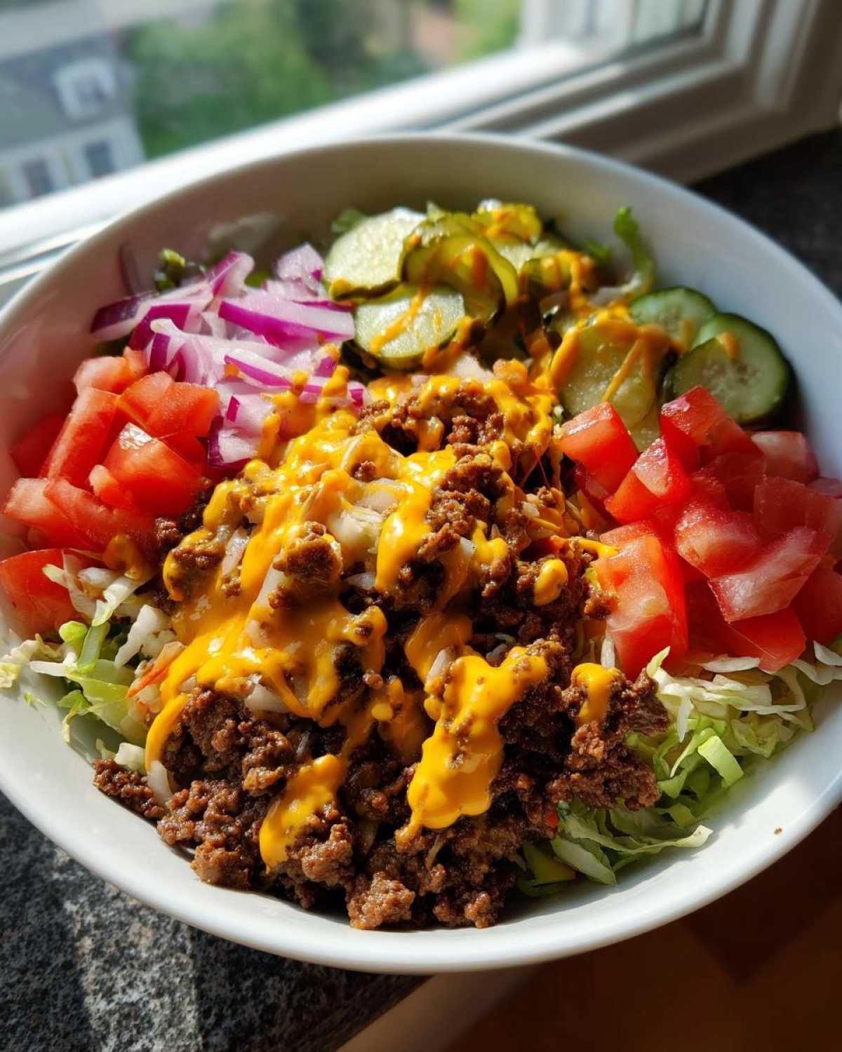 A close-up of a high-protein cheeseburger bowl featuring seasoned ground beef, melted cheese sauce, lettuce, tomatoes, red onion, and pickles.