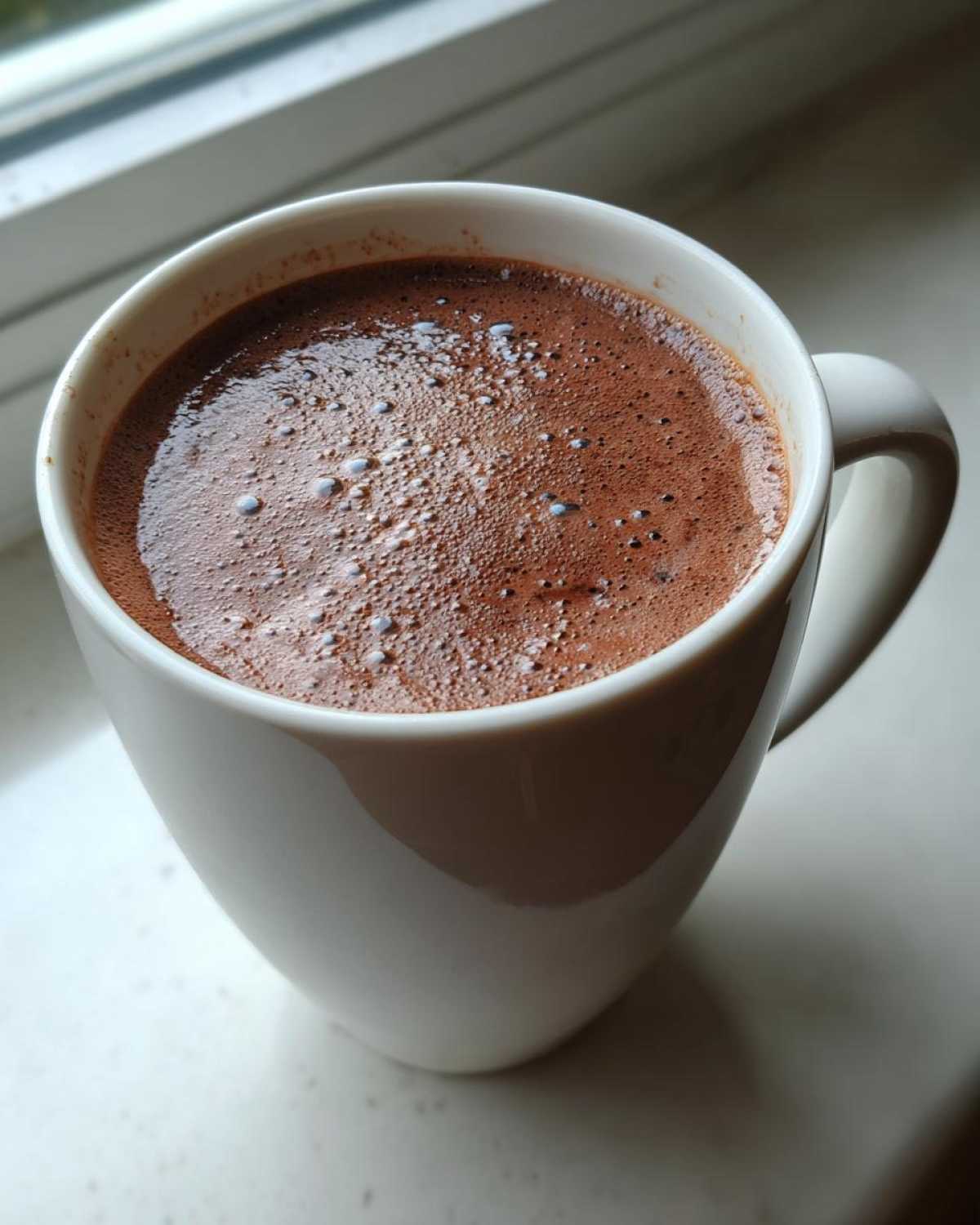 Close-up of rich, foamy hot cocoa mix in a white ceramic mug resting on a windowsill.