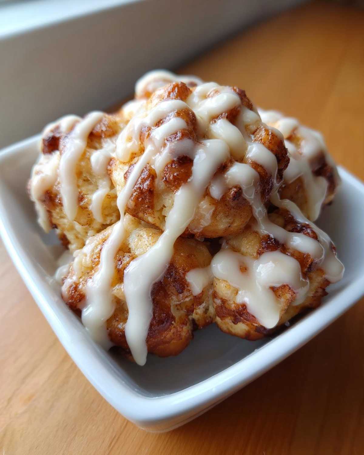 A close-up of several warm cinnamon roll bites drizzled generously with white icing, served in a small white square bowl.