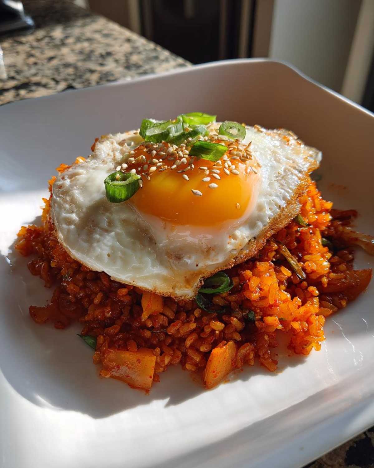 A serving of vibrant red kimchi fried rice topped with a sunny-side-up egg, sesame seeds, and green onions.