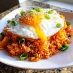 A close-up of vibrant kimchi fried rice topped with a sunny-side-up egg, sesame seeds, and green onions.