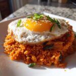 A mound of vibrant orange kimchi fried rice topped with a sunny-side-up egg, sesame seeds, and sliced green onions.