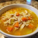 Close-up of a white bowl filled with vibrant Lemon Ginger Turmeric Chicken and Rice Soup, showing shredded chicken, rice, carrots, and celery.