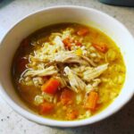Close-up of a white bowl filled with vibrant Lemon Ginger Turmeric Chicken and Rice Soup, topped with shredded chicken.