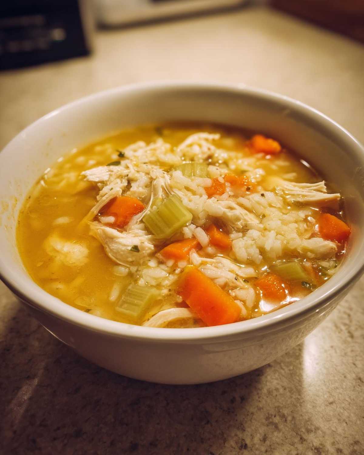 Close-up of a white bowl filled with rich, yellow Lemon Ginger Turmeric Chicken and Rice Soup, featuring shredded chicken, rice, carrots, and celery.
