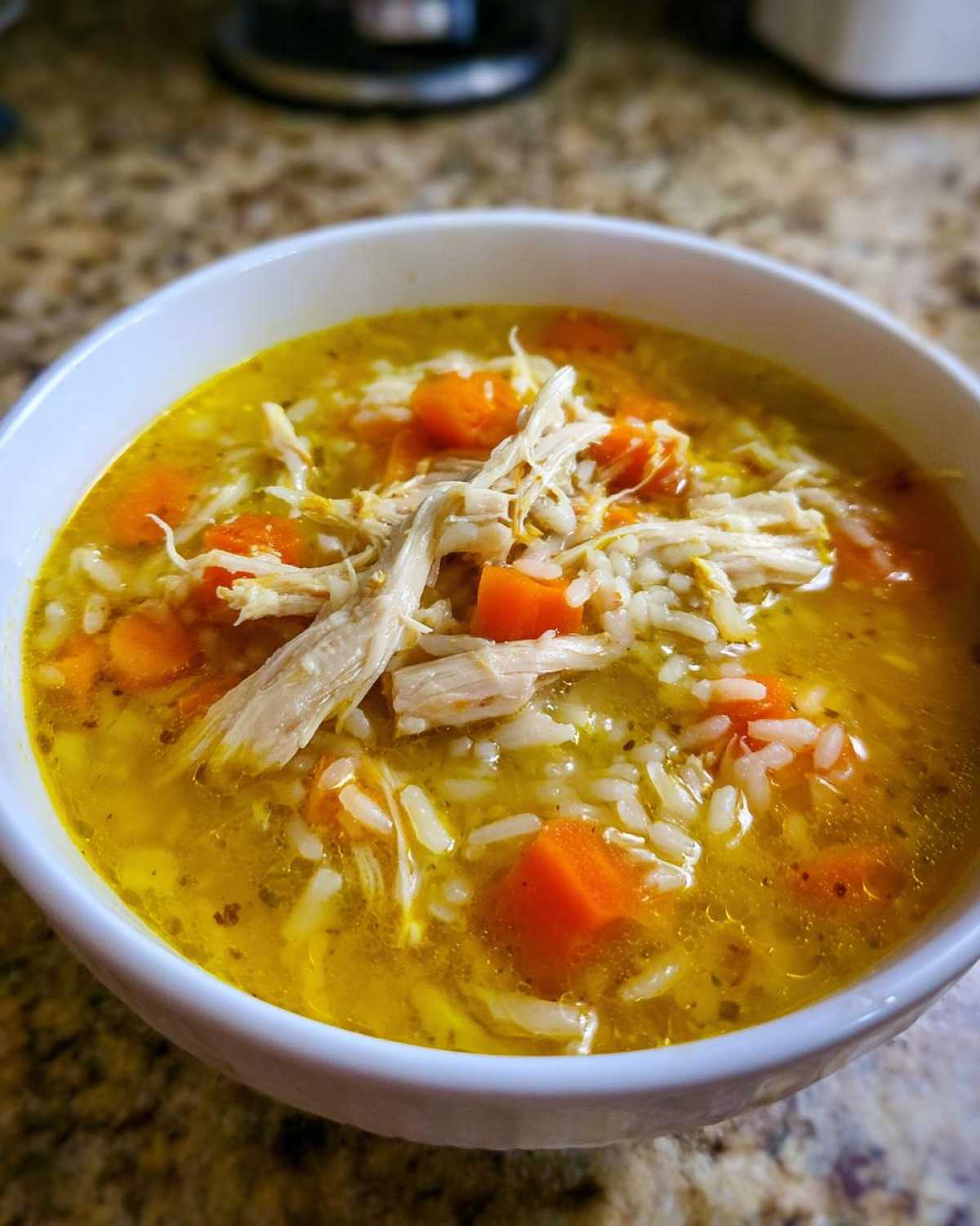 Close-up of a white bowl filled with vibrant yellow Lemon Ginger Turmeric Chicken and Rice Soup, featuring shredded chicken, rice, and diced carrots.