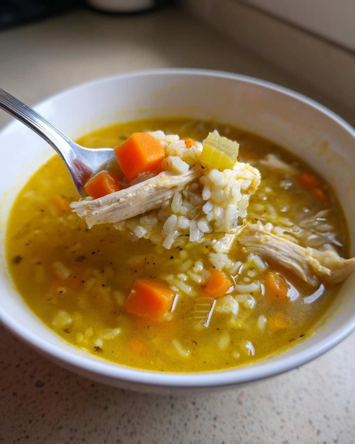 A spoonful of bright yellow Lemon Ginger Turmeric Chicken and Rice Soup showing shredded chicken, rice, and diced carrots.