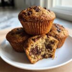 A stack of three moist banana chocolate chip muffins, one broken open showing melted chocolate chunks inside.