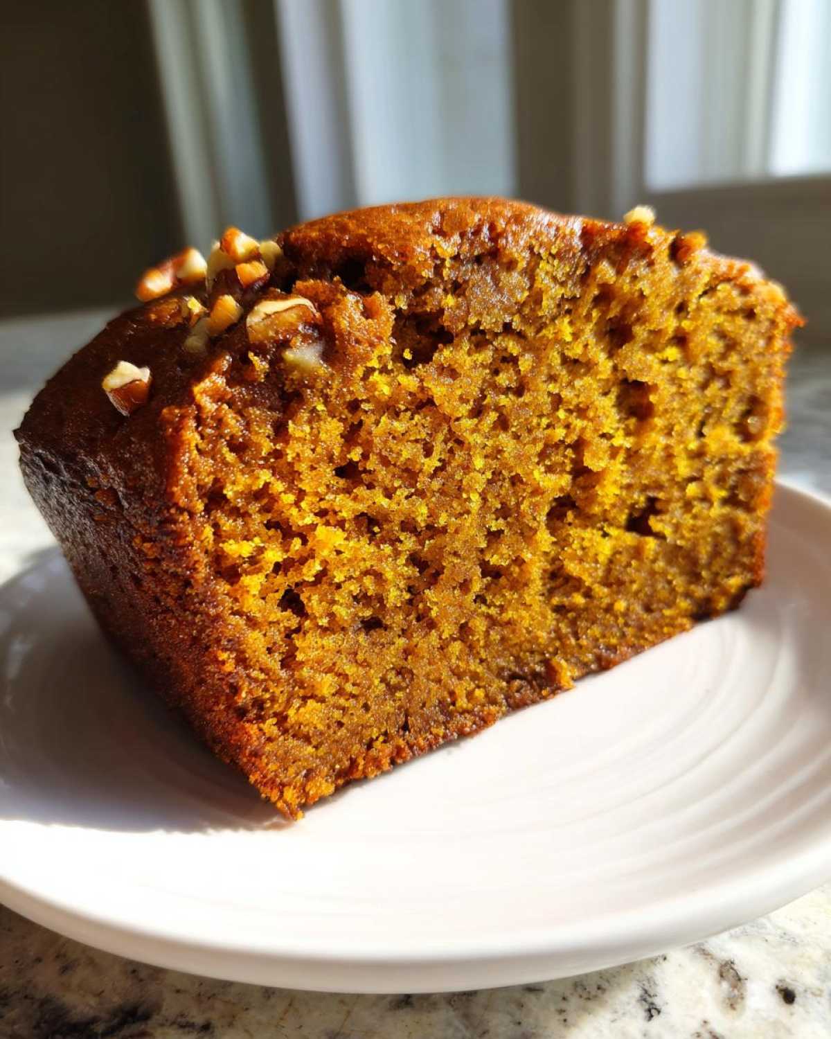 Close-up of a moist, orange-colored slice of pumpkin bread topped with chopped pecans on a white plate.