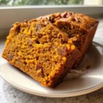 Two slices of moist, orange-colored pumpkin bread with nuts and raisins on a white plate.