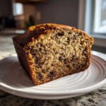 A close-up, sunlit slice of moist vegan banana bread packed with chocolate chips, resting on a white plate.