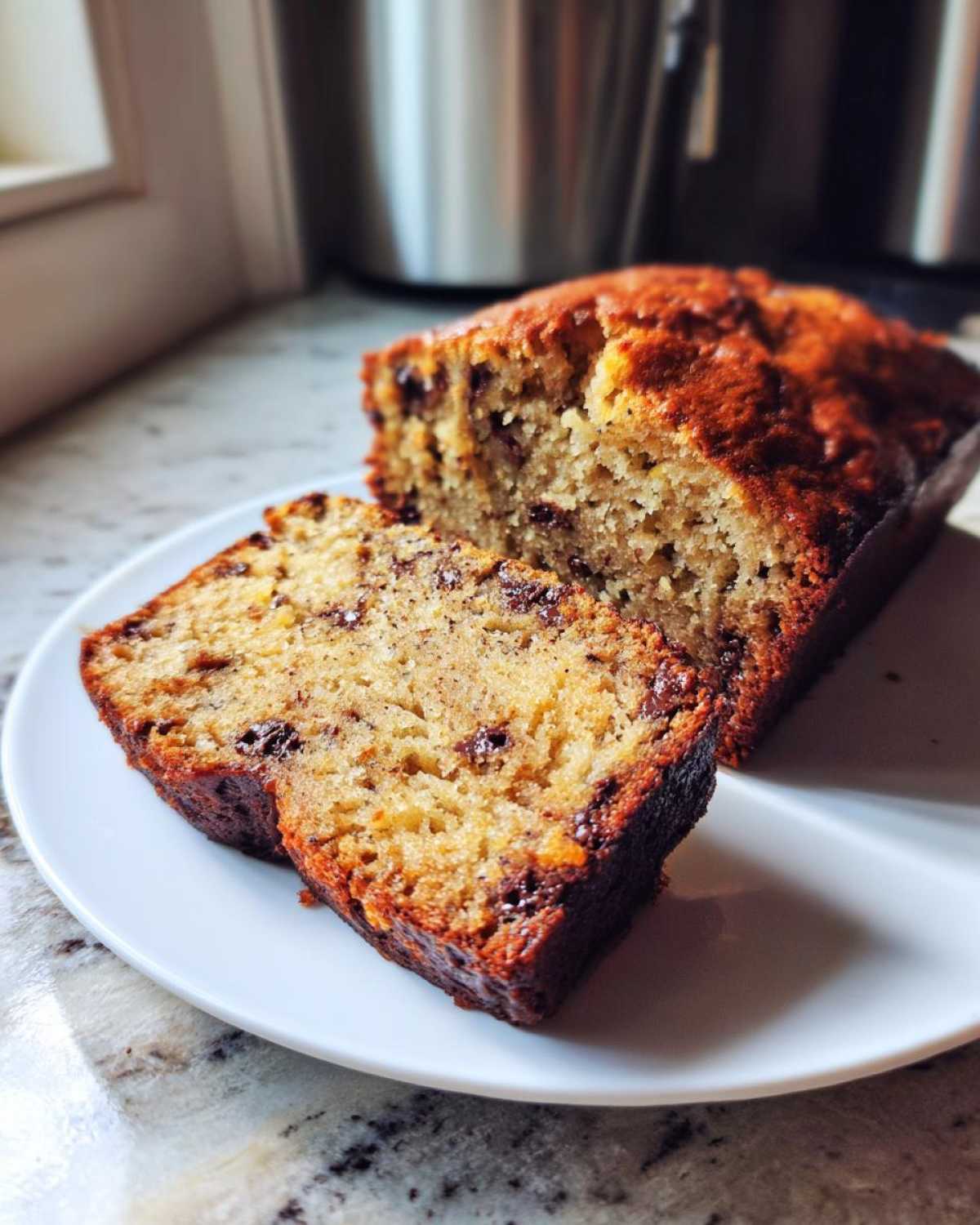 Close-up of moist vegan banana bread, sliced and showing chocolate chips, resting on a white plate.