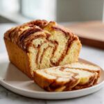 A freshly baked loaf of cinnamon bread, partially sliced, showing beautiful swirls of cinnamon filling.