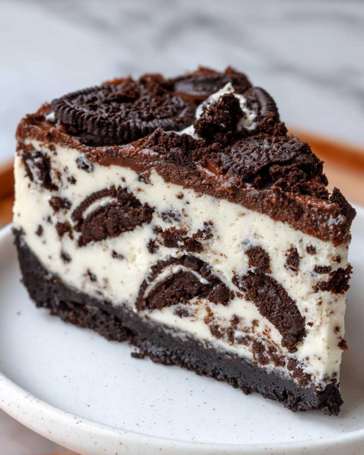 A perfect slice of Oreo cheesecake showing a creamy white filling loaded with crushed Oreo pieces and topped with chocolate ganache and whole cookies.