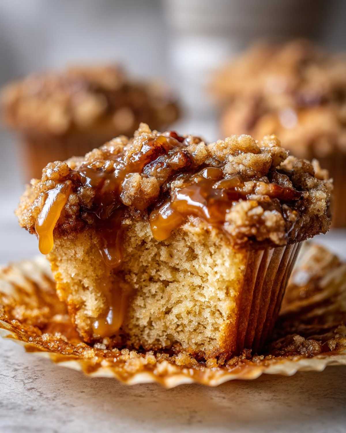 A close-up of a pecan pie muffin with a bite taken out, showing the crumb texture and gooey caramel dripping over the streusel topping.
