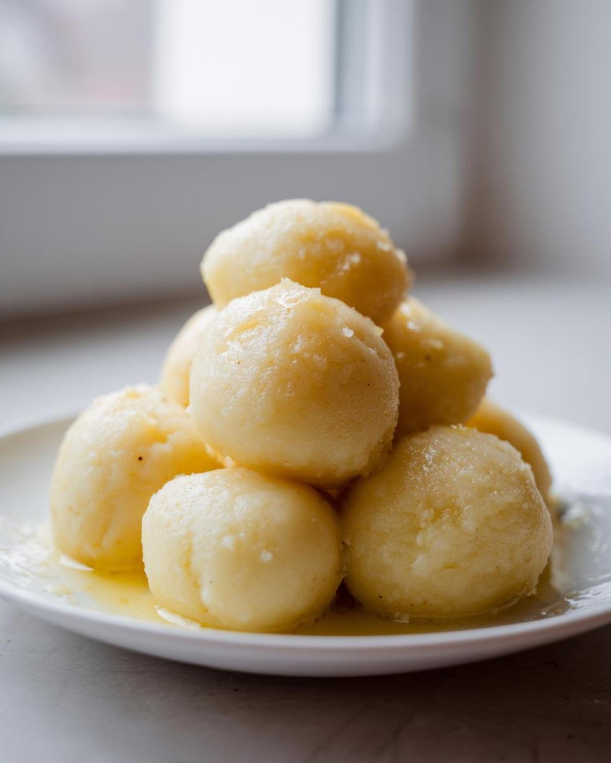 A stack of freshly made, glistening potato dumplings served in melted butter on a white plate.