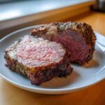 Two thick slices of medium-rare prime rib recipe roast with a dark herb crust on a white plate.