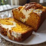A loaf of bright orange pumpkin cream cheese bread, partially sliced, showing the creamy white swirl filling.