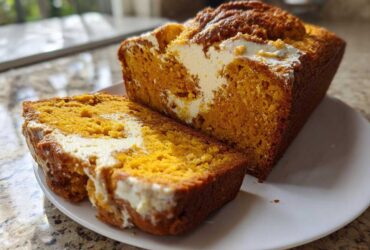 A loaf of bright orange pumpkin cream cheese bread, partially sliced, showing the creamy white swirl filling.