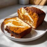 A loaf of rich orange pumpkin cream cheese bread, partially sliced, showing a beautiful cream cheese swirl inside.