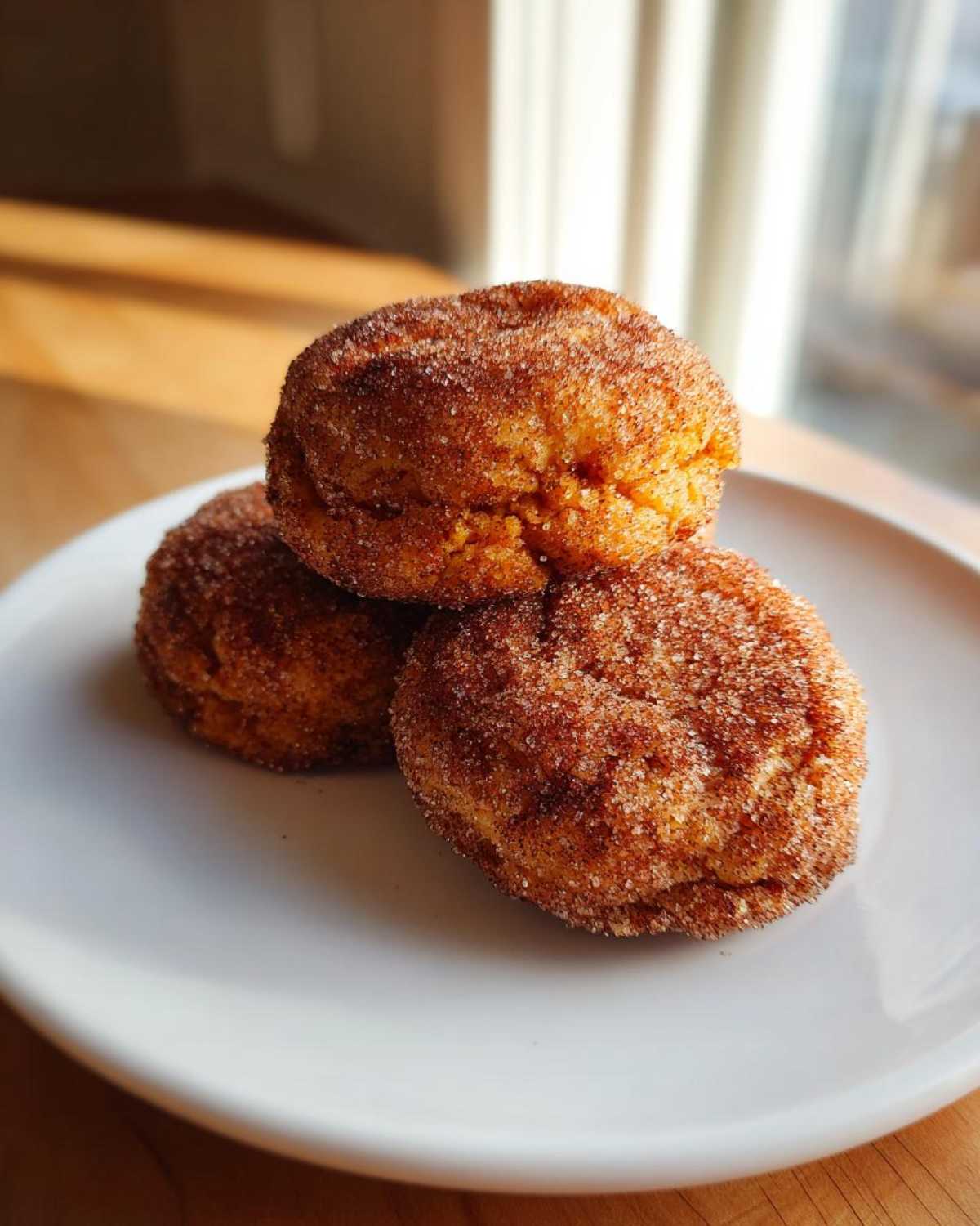 Three freshly baked pumpkin snickerdoodle cookies heavily coated in cinnamon sugar, stacked on a white plate.