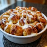 A white bowl filled with freshly baked, golden brown cinnamon roll bites drizzled generously with white icing.