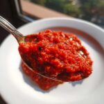 Close-up of a spoonful of thick, vibrant red pizza sauce with visible herbs, held over a white plate.