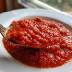 Close-up of a spoonful of thick, vibrant red pizza sauce, showing herbs, over a white bowl.