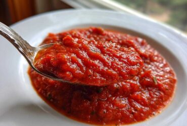 Close-up of a spoonful of thick, vibrant red pizza sauce, showing herbs, over a white bowl.