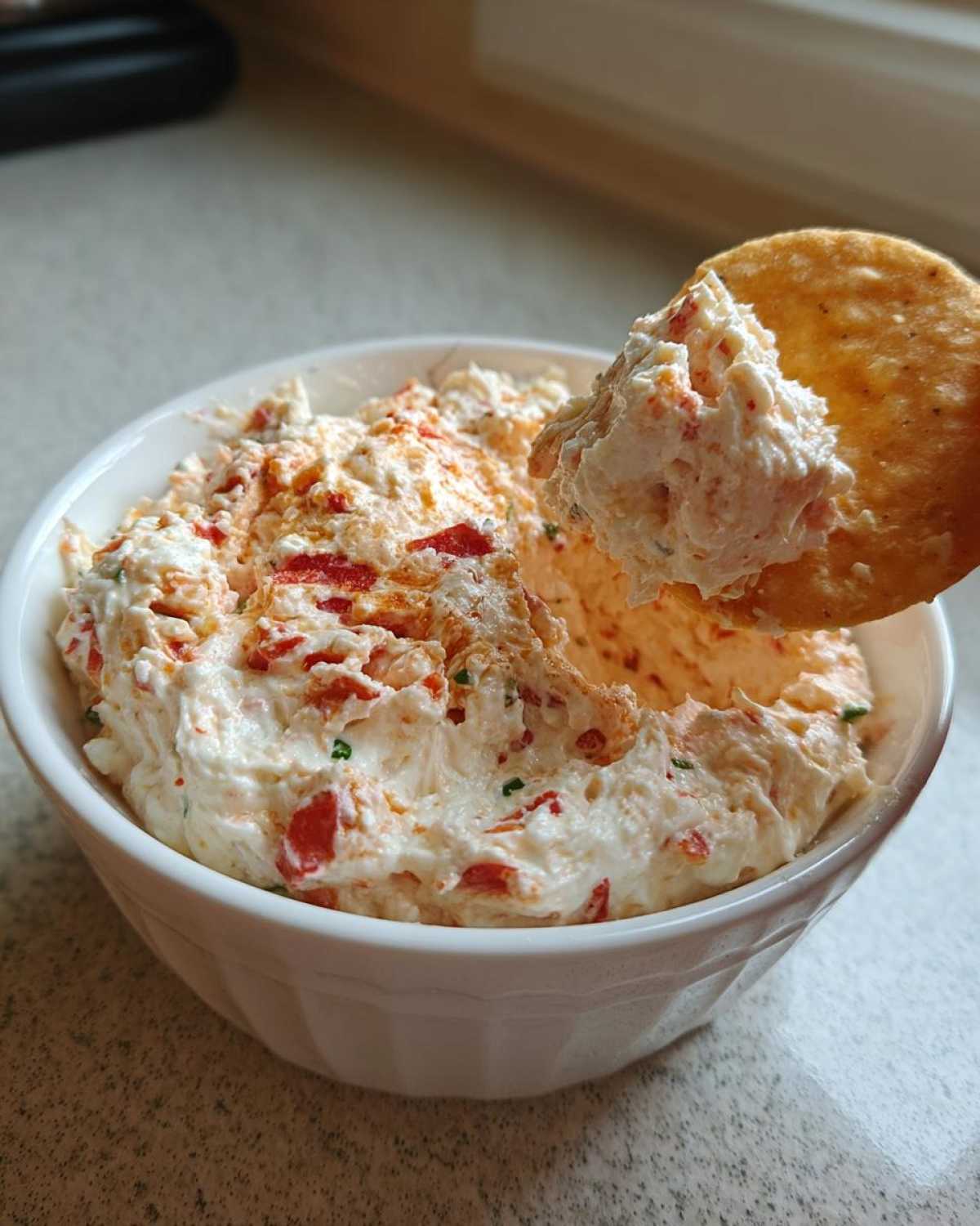 A cracker scooping a generous amount of creamy, orange-flecked boat dip from a white bowl.