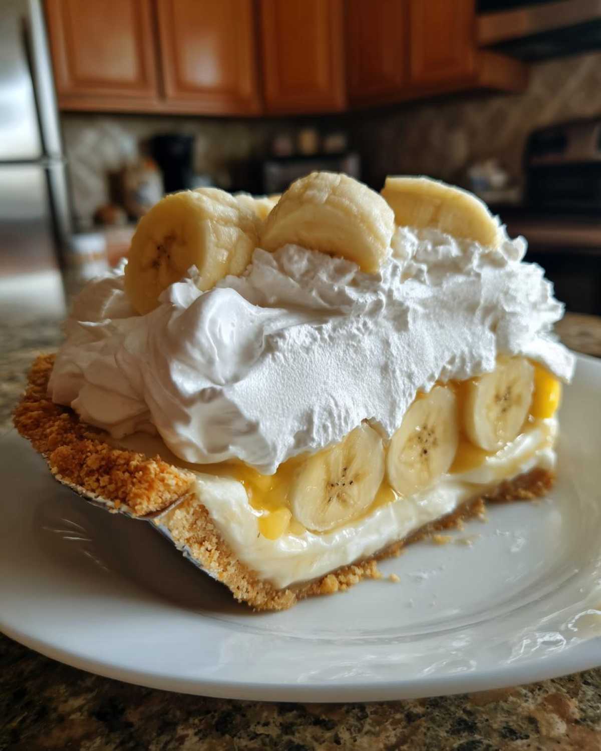 A generous slice of banana cream pie with a graham cracker crust, layered with bananas, cream filling, and topped with whipped cream.