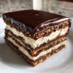 A close-up of a decadent slice of chocolate eclair cake showing multiple layers of graham crackers, cream filling, and rich chocolate ganache.