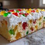 Close-up of a slice of moist jello poke cake showing colorful gelatin pieces inside the vanilla cake, topped with whipped cream and candied fruit.