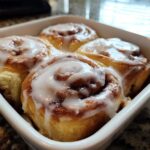 Four freshly baked small batch cinnamon rolls drizzled with thick white icing in a white square baking dish.