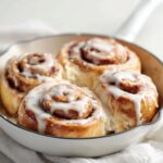 Four gooey small batch cinnamon rolls topped with white icing baked in a white cast iron skillet.