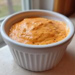 Close-up of a creamy, orange-hued smash burger sauce speckled with spices, served in a small white fluted ramekin.