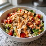 A white bowl filled with southwest chicken salad, featuring shredded chicken, tomatoes, black beans, and a creamy orange dressing.