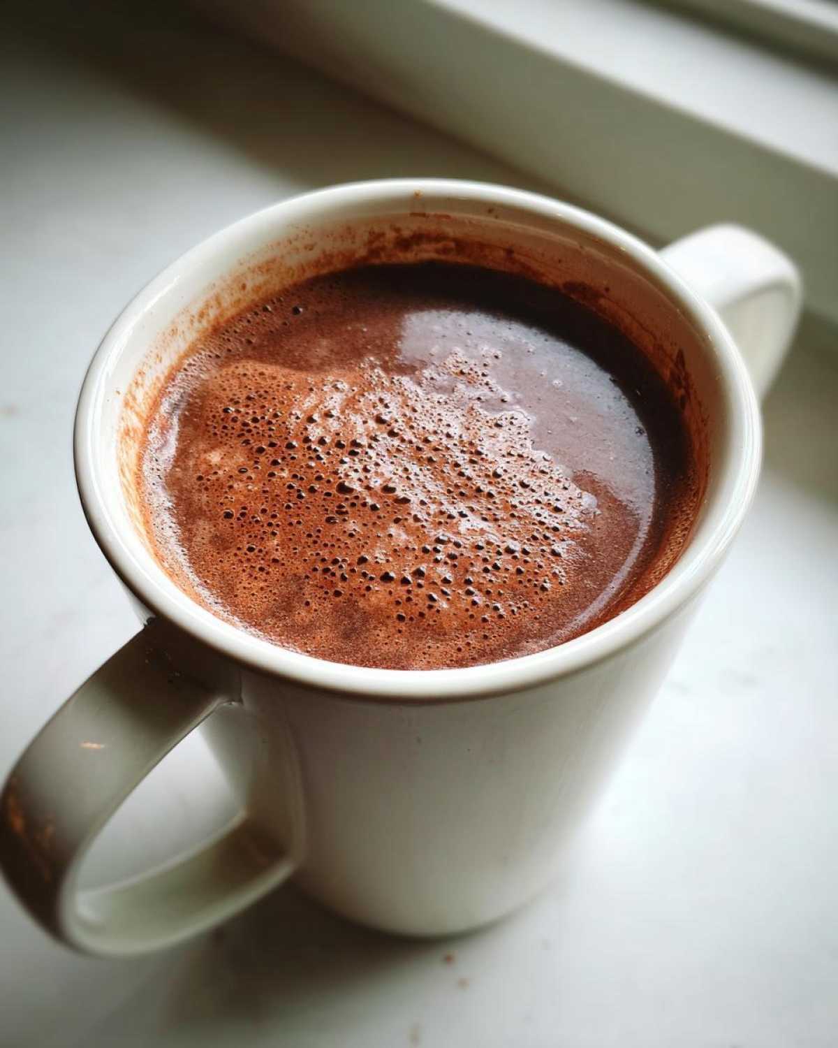 Close-up of a white mug filled with rich, frothy hot cocoa mix sitting on a bright windowsill.