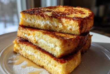Three thick slices of golden brown, stuffed french toast stacked on a white plate, drizzled with syrup.