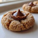 A close-up of a freshly baked, sugar-coated peanut butter kiss cookie with a melted chocolate kiss pressed into the center.