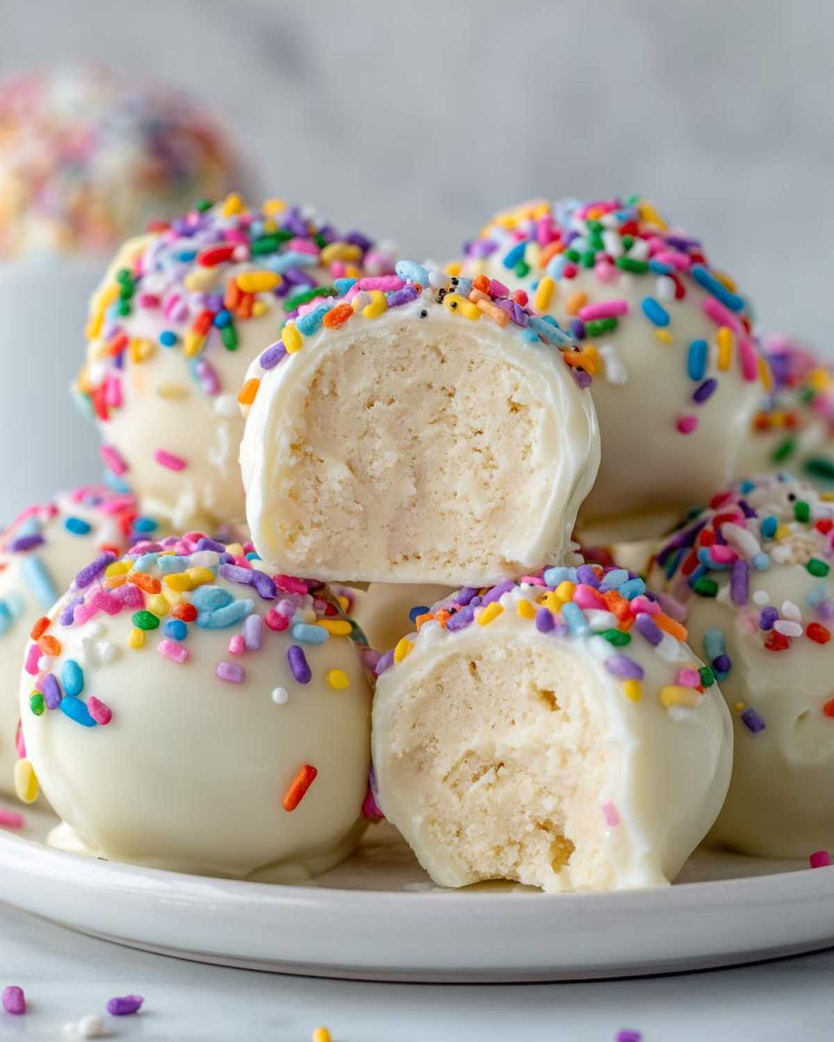 A stack of white chocolate-coated sugar cookie truffles covered in colorful sprinkles, one cut in half showing the creamy interior.